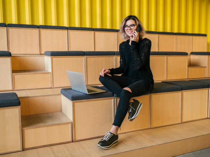 young attractive woman sitting in lecture hall, working on laptop, wearing glasses, modern auditorium, student education online, freelancer, smiling, talking on phone