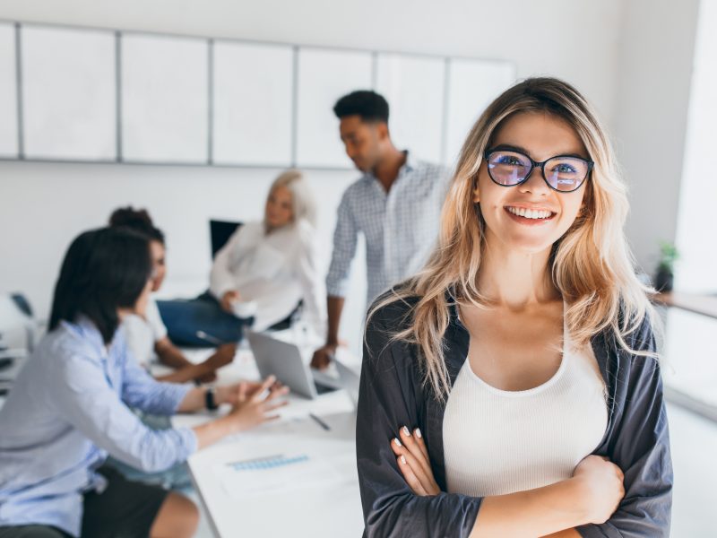 Blonde female executive posing with smile and arms crossed during brainstorm with managers. Indoor portrait of european student spending time in hall with asian and african friends.