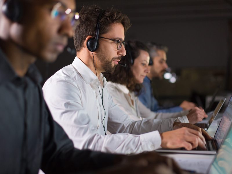 Side view of confident call center operator talking with client. Caucasian young man in eyeglasses typing on laptop while serving client. Call center concept