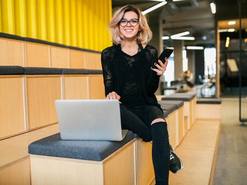 young attractive woman sitting in lecture hall, working on laptop, wearing glasses, modern auditorium, student education online, freelancer, smiling, using smartphone, looking in camera