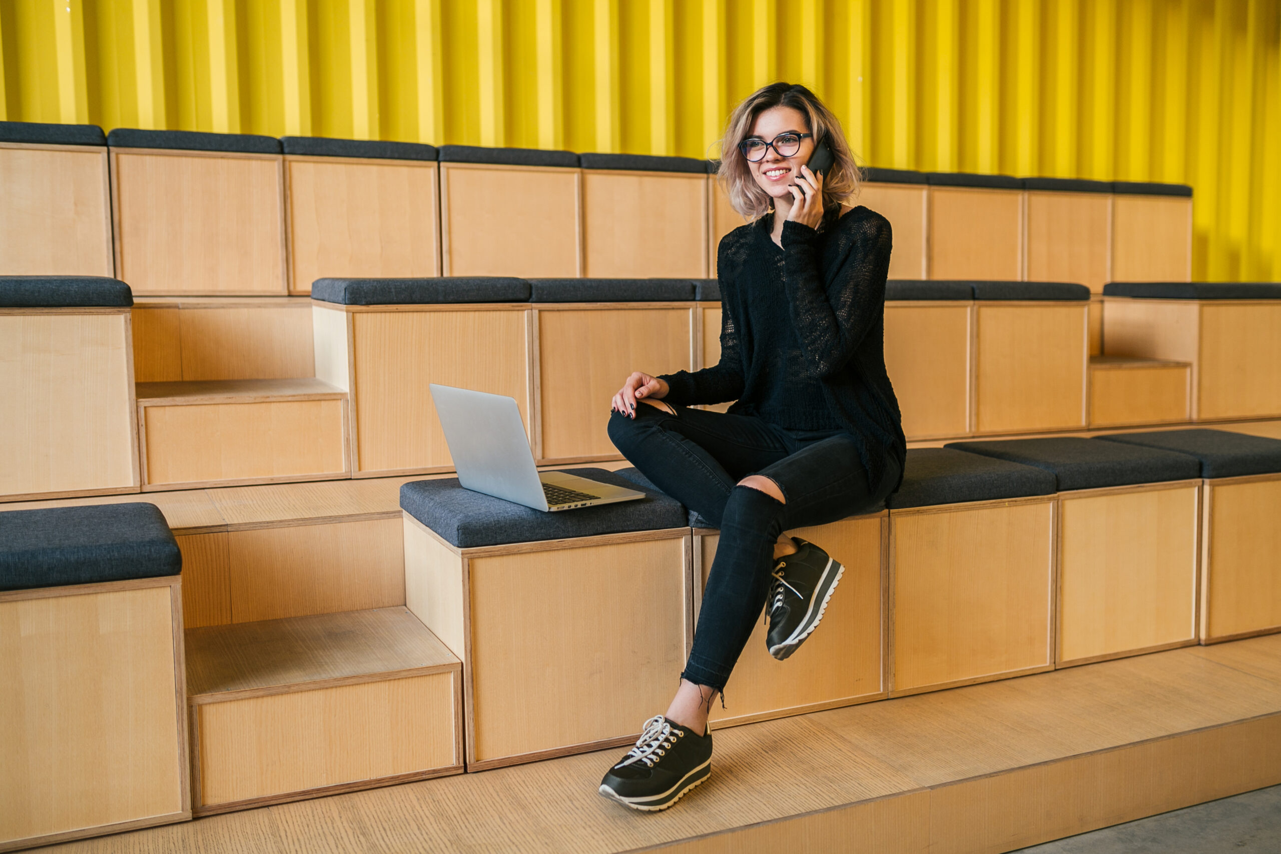 young attractive woman sitting in lecture hall, working on laptop, wearing glasses, modern auditorium, student education online, freelancer, smiling, talking on phone