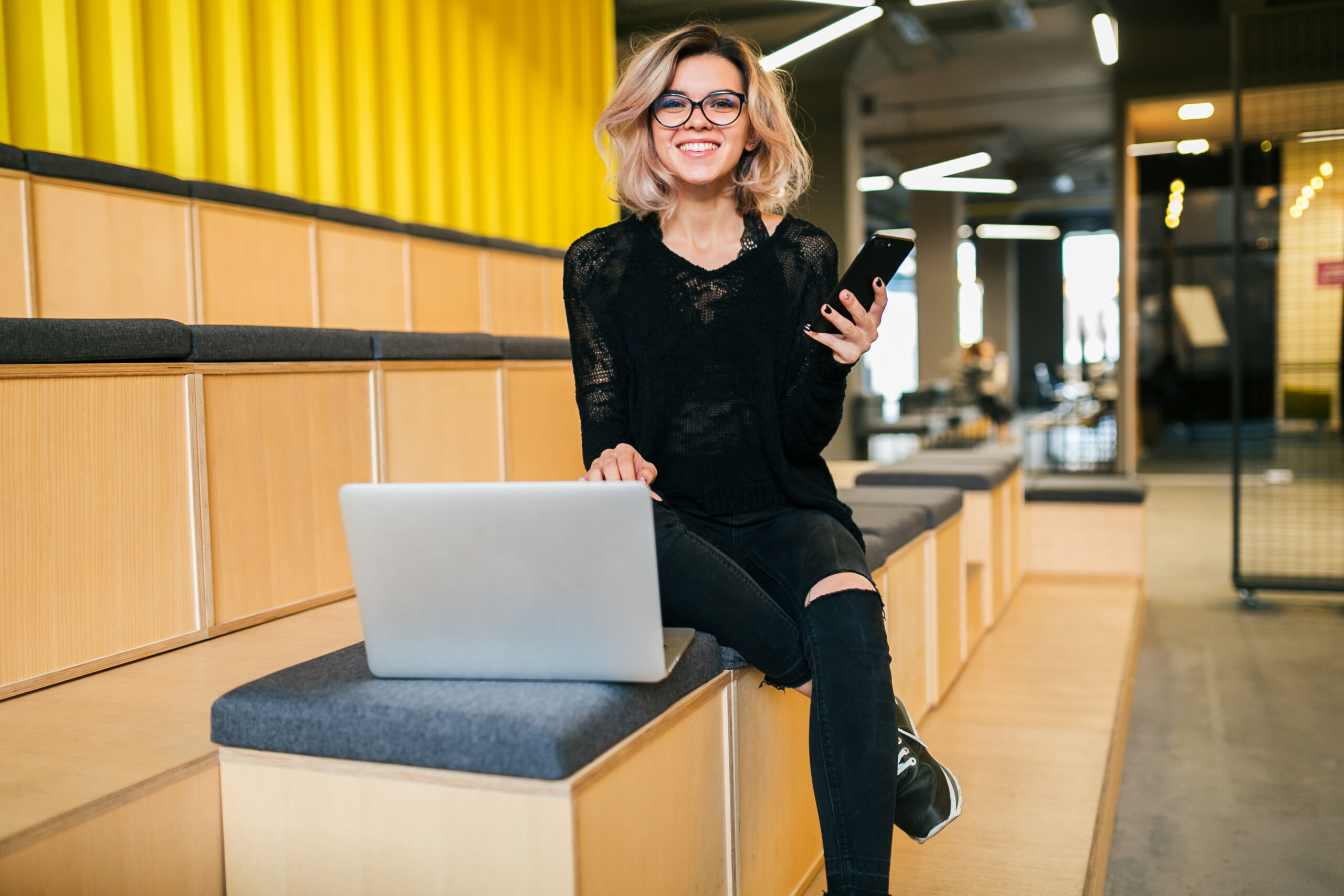 young attractive woman sitting in lecture hall, working on laptop, wearing glasses, modern auditorium, student education online, freelancer, smiling, using smartphone, looking in camera